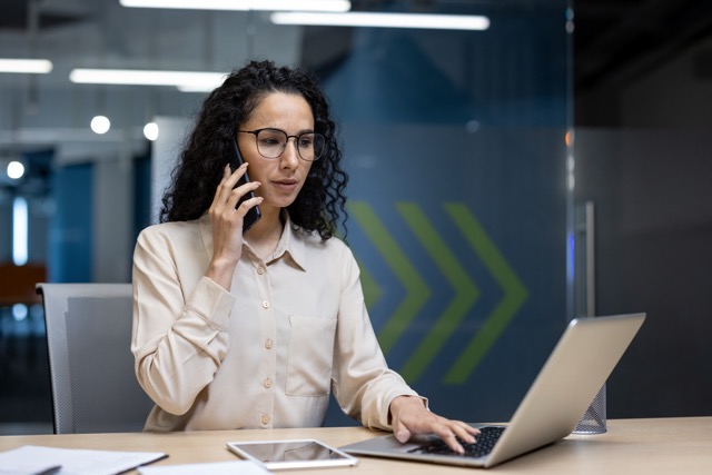 A woman is talking on the phone and working on her laptop
