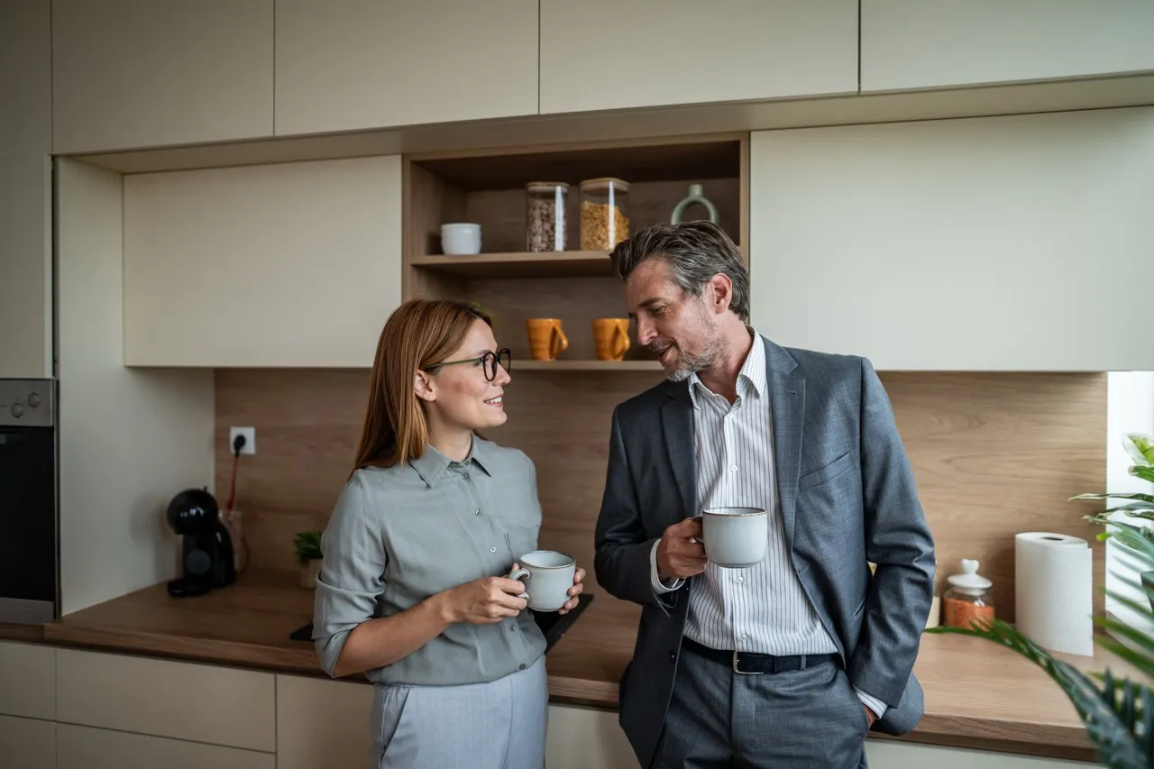 An elderly couple drinks coffee and chats in the kitchen.