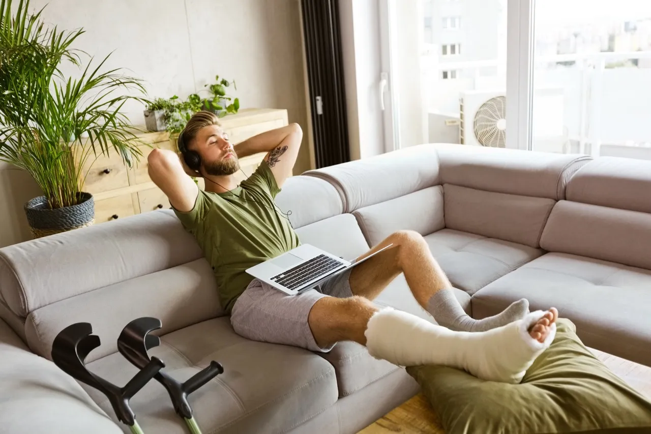 A man with a broken leg is sitting on a sofa, holding a laptop and thinking
