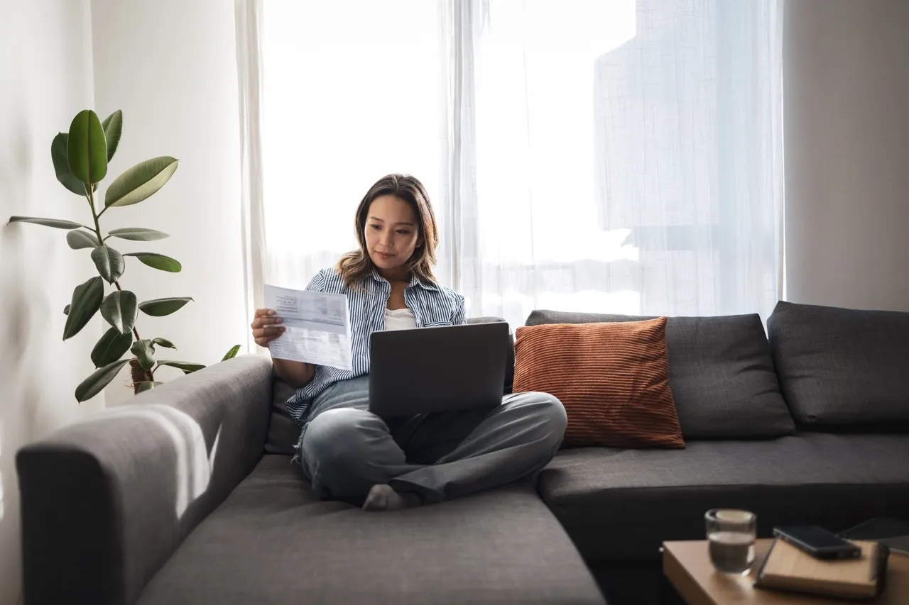 Woman sits on sofa with laptop and checks invoice